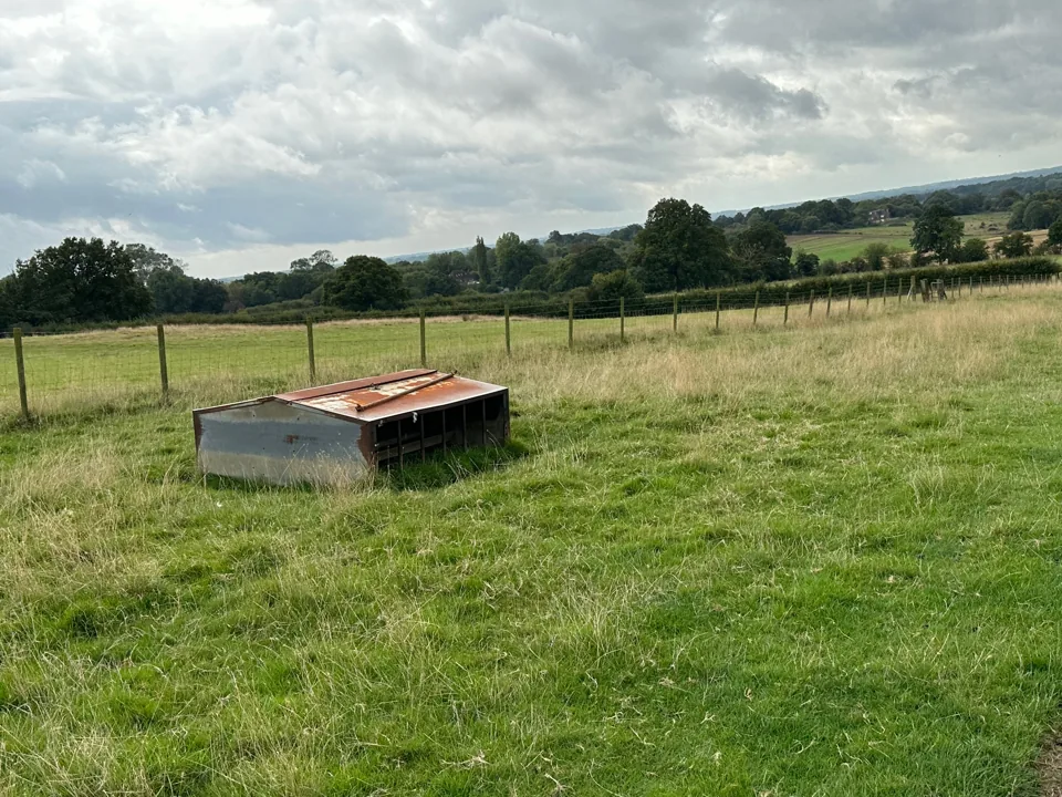 Fenced pasture with a water trough and wooded countryside beyond.