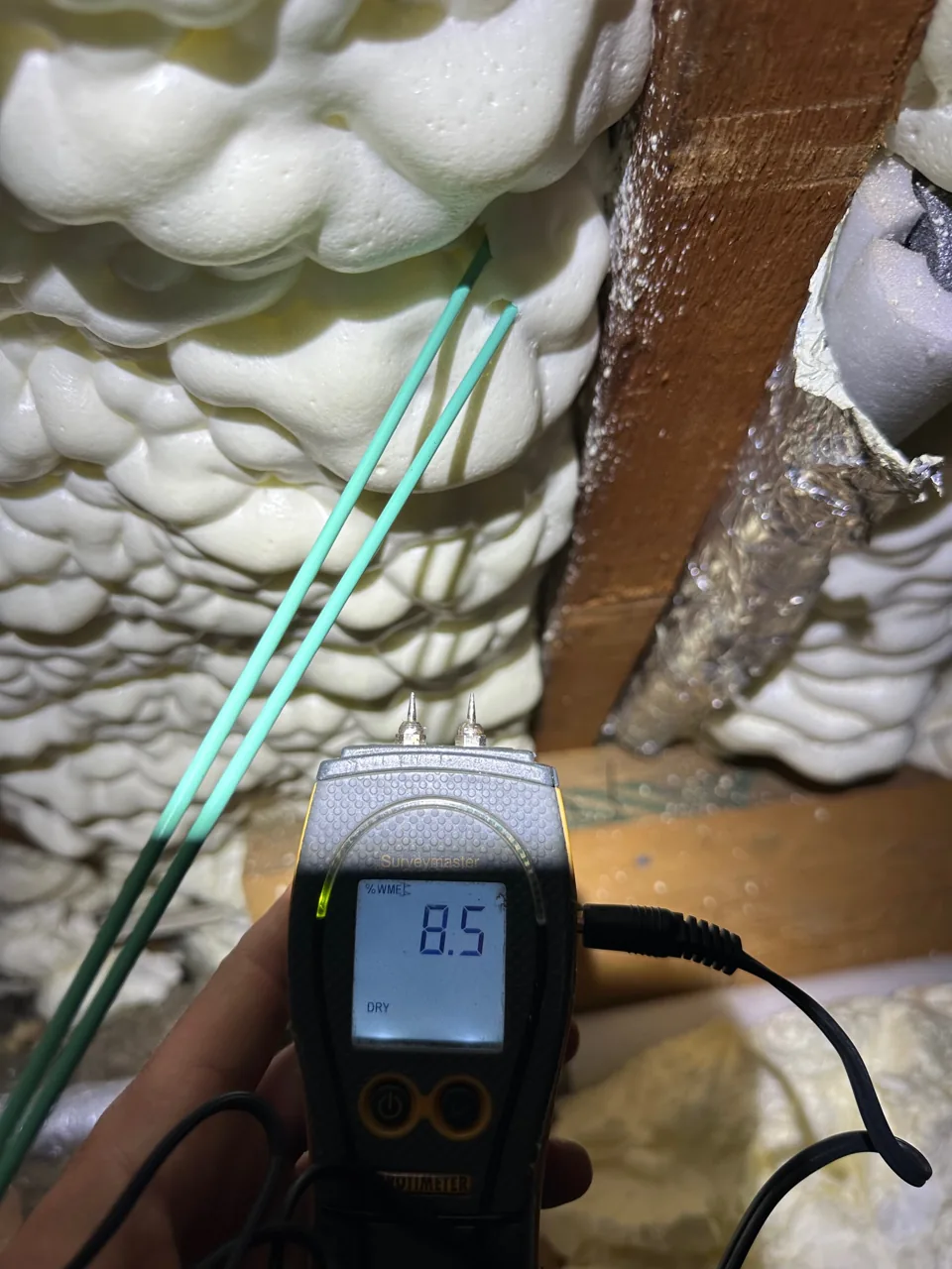 A handheld moisture meter beside a roof timber and foam insulation.