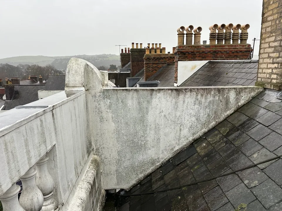 Sloping slate roof beside a rendered parapet, with chimney stacks beyond.